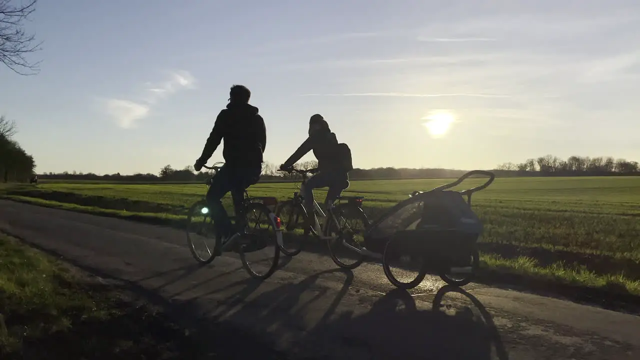 Slow motion shot showing silhouette of family on bikes with child transporter riding on rural road during sunset