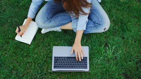 Woman student working on laptop computer and writing notes in notebook in park