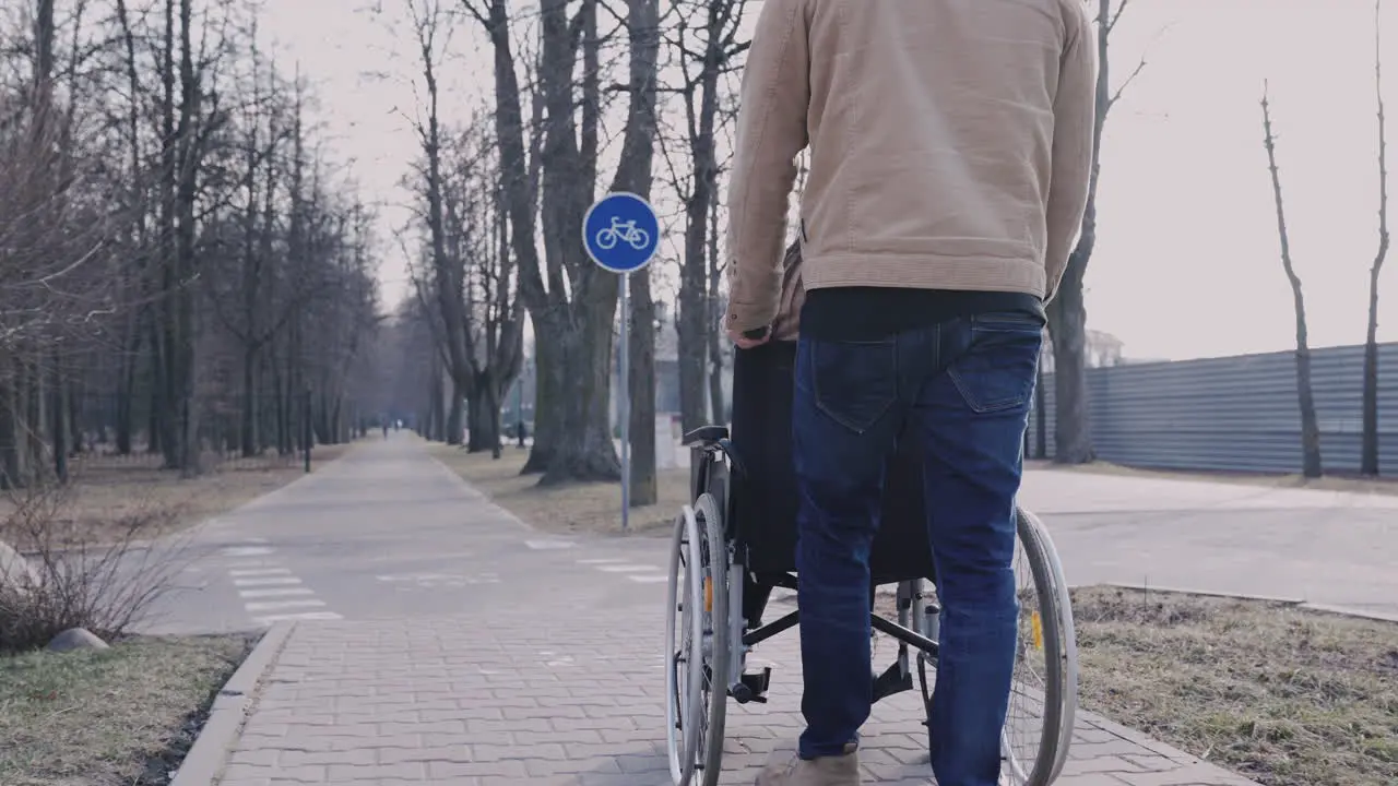 Rear View Of A Man Taking Her Disabled Friend In Wheelchair On A Walk Around The City