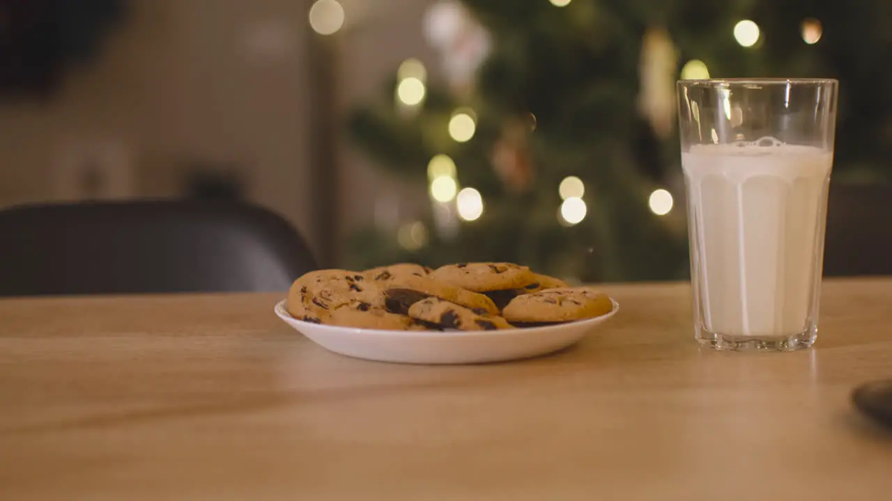 Camera Focuses On An Glass Of Milk And A Plate Full Of Cookies On An Empty Table In A Room Decorated With A Christmas Tree