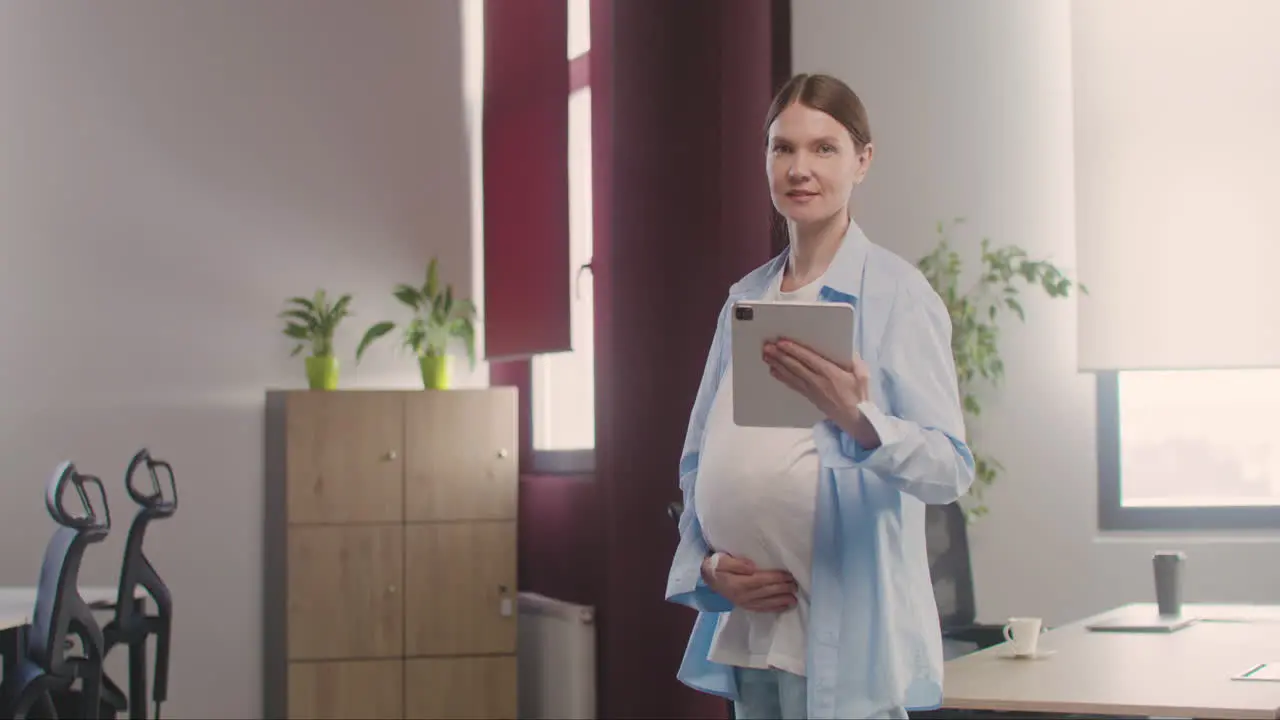 Pregnant Woman Posing At Camera While Touching Her Belly And Holding Tablet In The Office