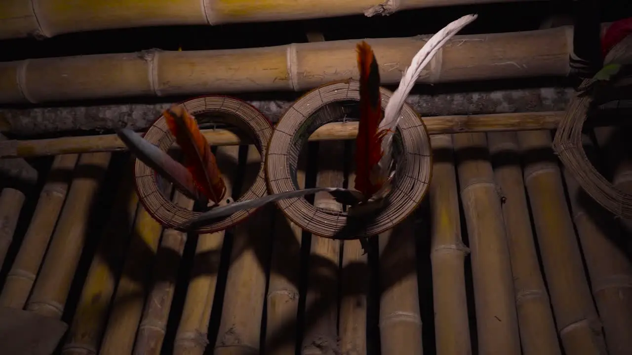 At night a cinematic slow-motion shot pans from right to left focusing on traditional bird-feathered hats of Shipibo indigenous people hung on the wall of a bamboo hut