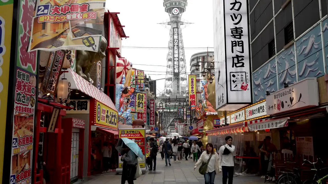 Visitors Exploring Shinsekai On Overcast Rainy Day With View Of Tsutenkaku Tower In The Background