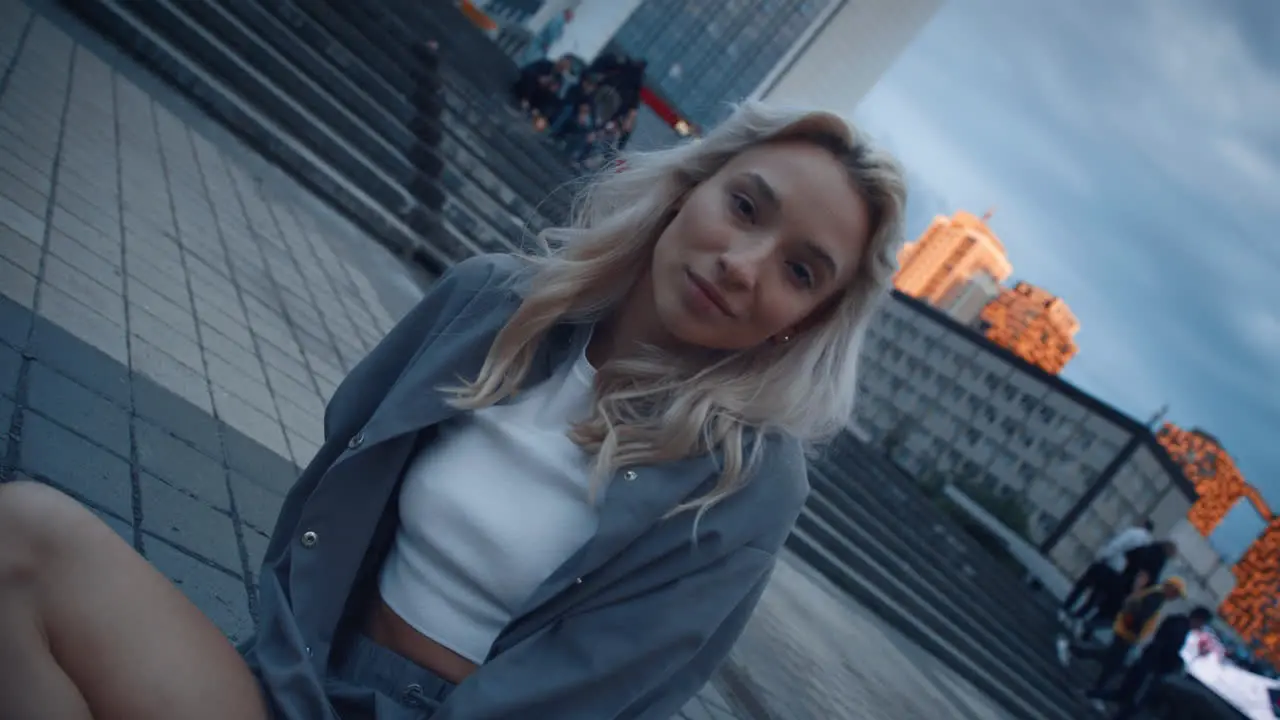 Smiling woman sitting city street near modern urban buildings at evening