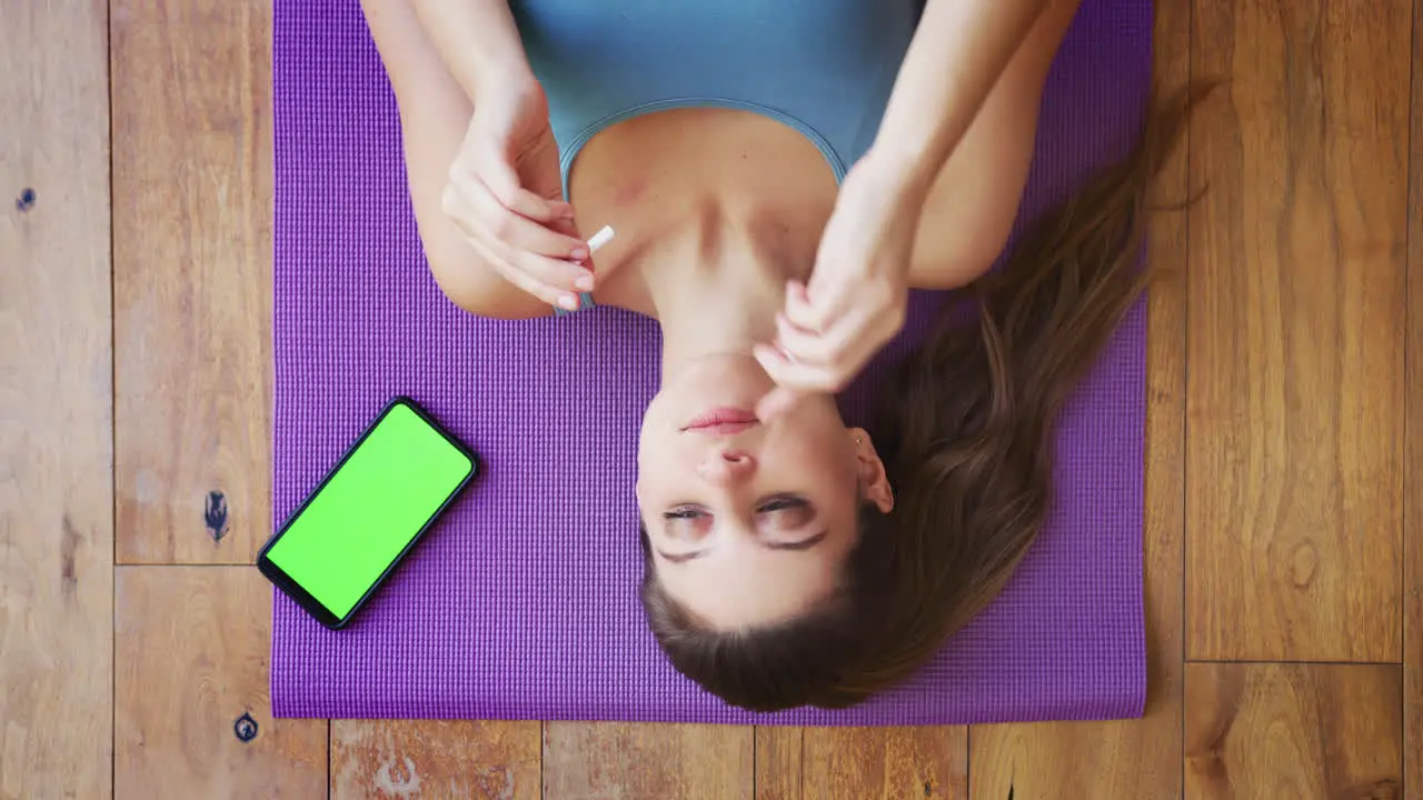 Overhead View Of Woman Lying On Exercise Mat At Home Putting In Wireless Earphones