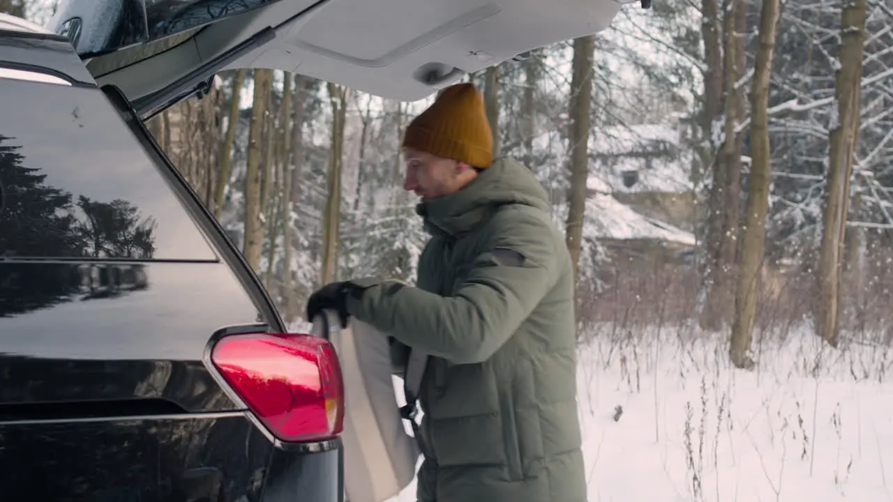 Side View Of A Man Dressed In Winter Clothes Putting Things Into The Trunk Of A Car In A Snowy Forest