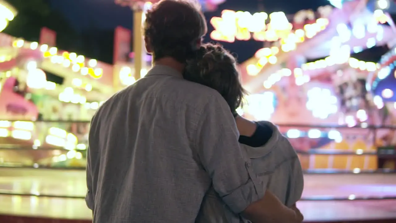 Young Couple Visiting An Amusement Park Arcade Together And Hugging While Standing Next To A Carousel Ride With Lights At Night