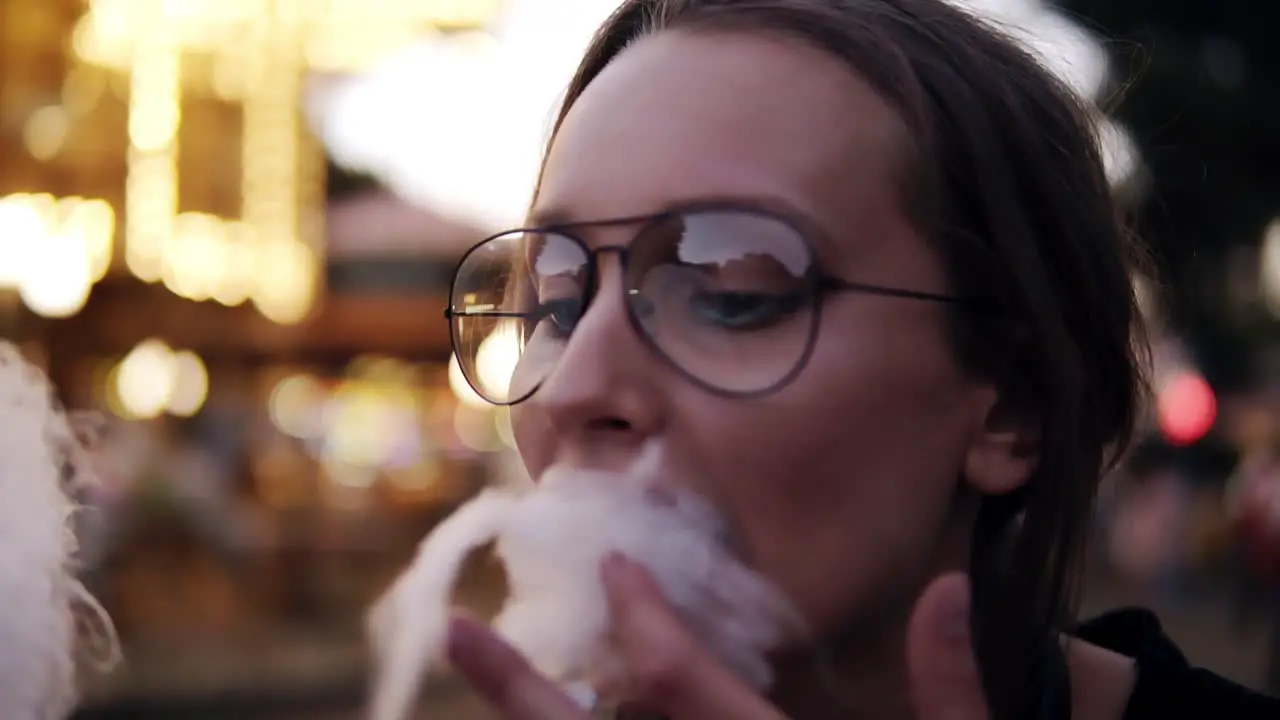 Beautiful Girl Eating Cotton Candy In An Amusement Park Near The Blurred Carousel