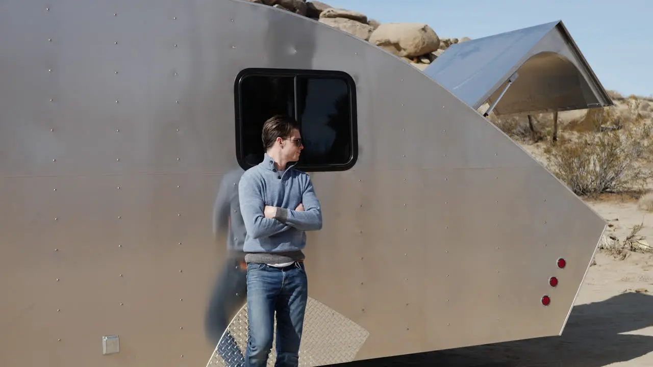 A handsome young white man traveler leaning against a metal teardrop travel trailer tiny house on his solo road trip