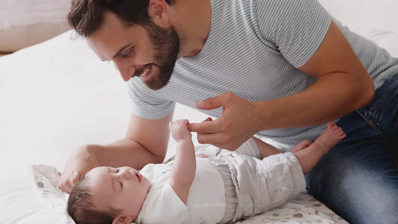 Close Up Of Loving Father Playing With Newborn Baby Son Lying On Bed At Home In Loft Apartment