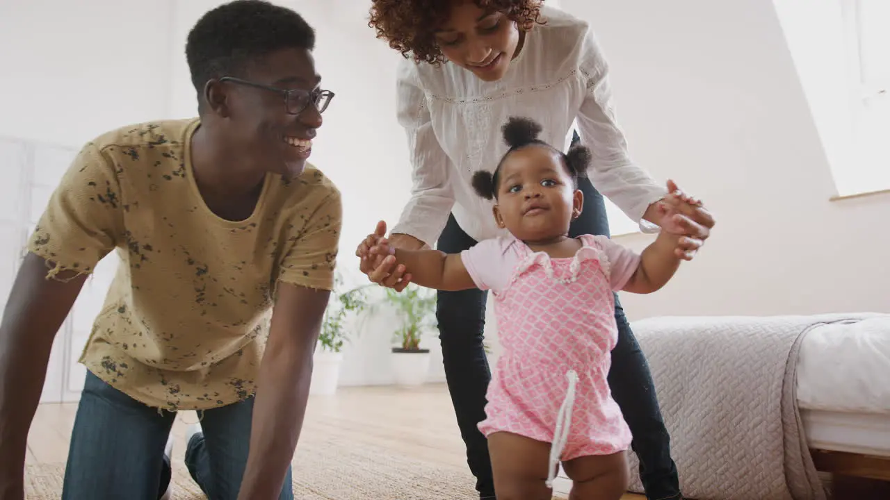 Baby Girl Learning To Walk At Home As Parents Sit On Floor And Give Encouragement