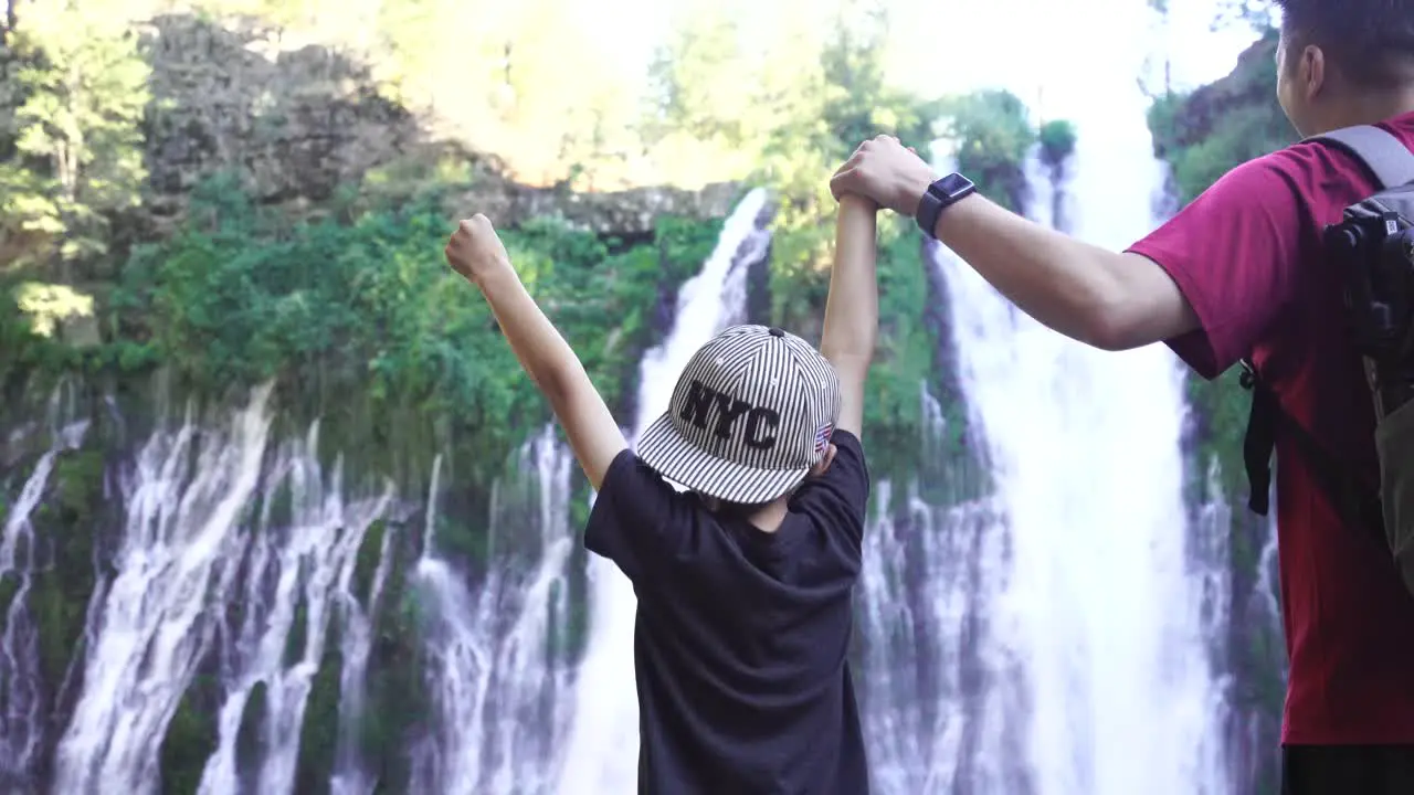 Father and son raising their hands together while holding hands in front of a gorgeous waterfall