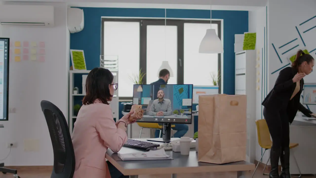 Entrepreneur woman sitting at desk in company office eating sandwich during online videocall conference