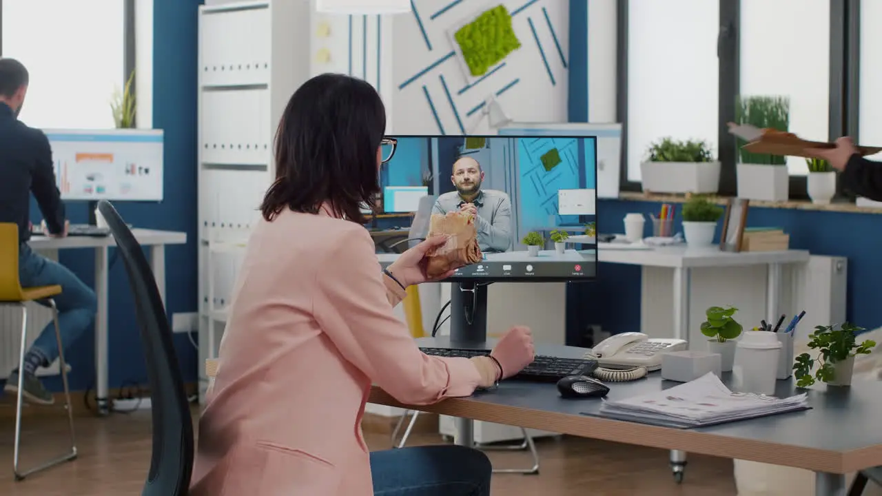 Businesswoman sitting at desk in company office eating sandwich during online videocall conference