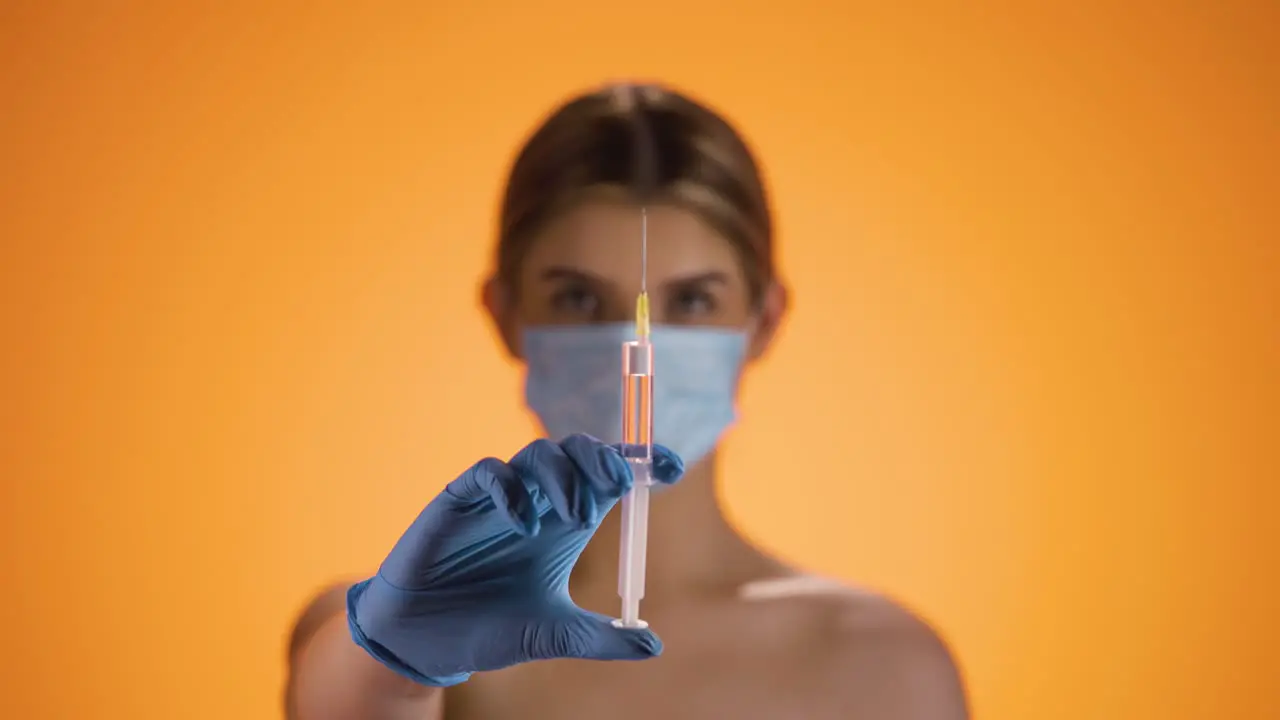 Medium shot of a young pretty nurse wearing a ffp2 mask while holding a syringe with orange liquid into the camera to vaccinate against diseases or corona virus in front of orange background