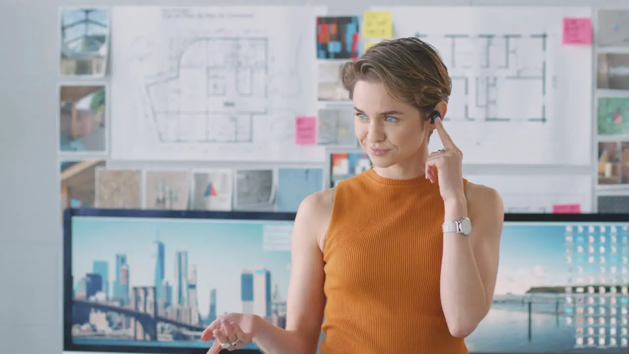 Female Architect Standing In Office Talking To Client Using Wireless Earphones