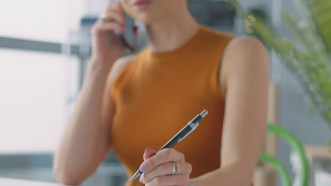 Female Architect In Office Working On Plans At Desk Whilst Talking On Mobile Phone