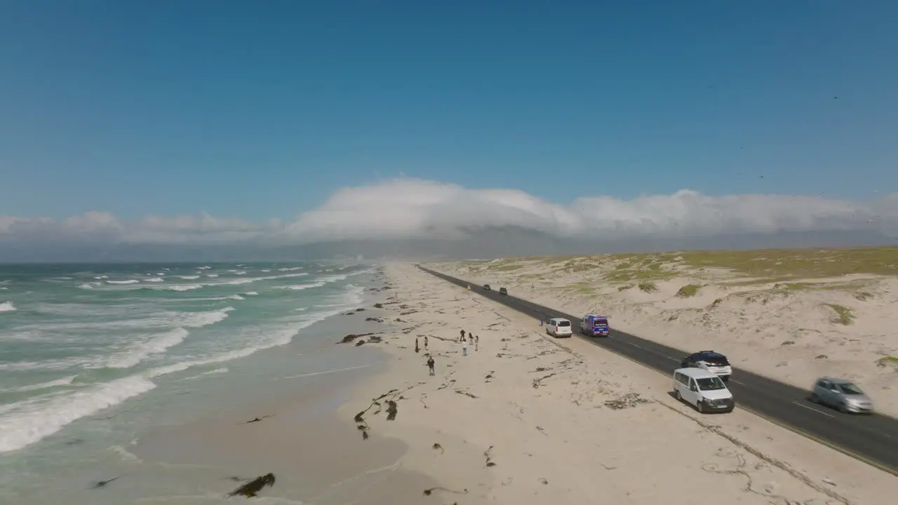 Cars driving on road along coast People taking rest and walking on sandy beach Waves washing shore