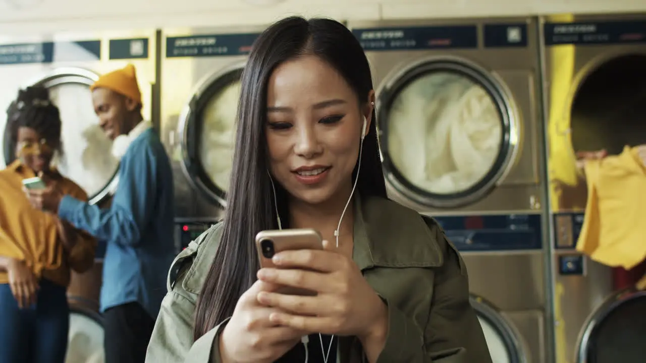 Pretty Cheerful Woman In Headphones Watching Video On Smarphone In Laundry Service Room