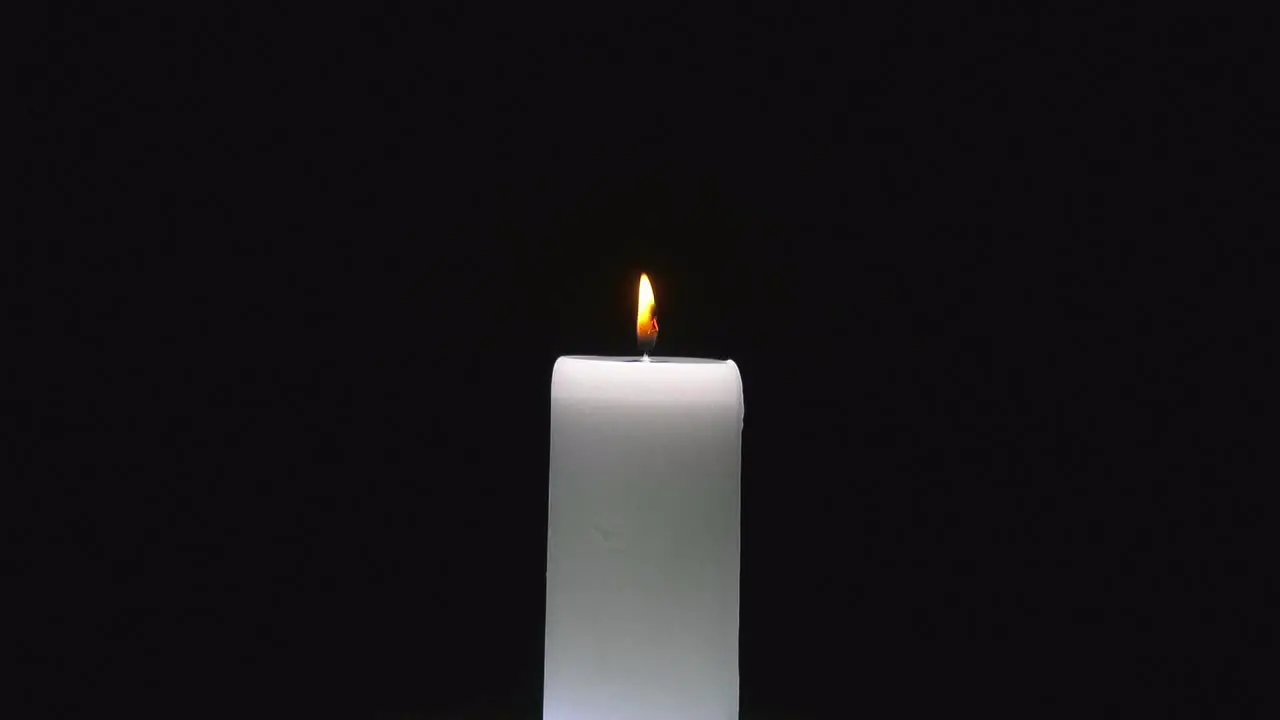 Small red flame flickers from white cylindrical candle rotating as wax pools and drips on black background