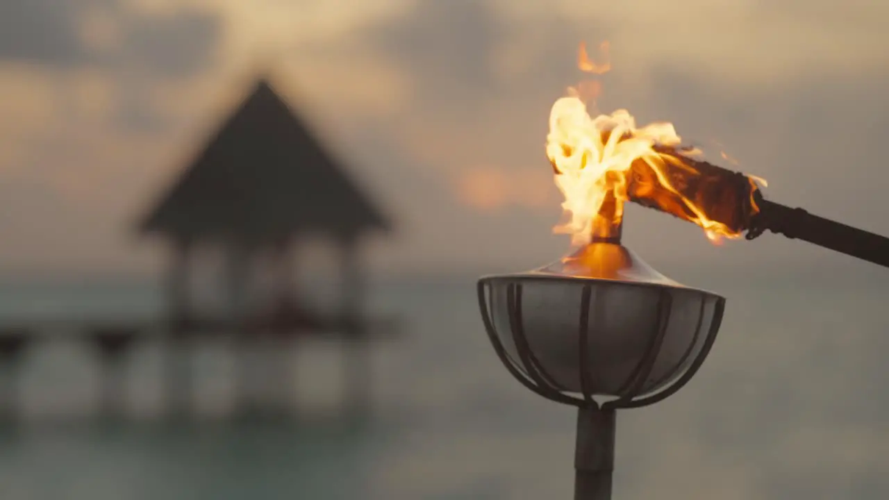 Steel oil lamp on post being lit by burning torch with dramatic sunset in background