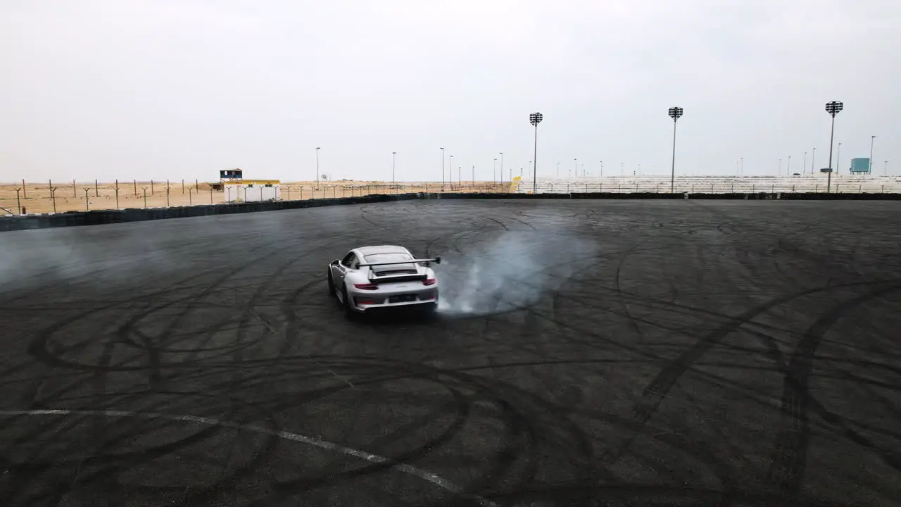 Aerial tracking shot of a grey Porsche GT3 RS doing donuts and burnouts in an arena