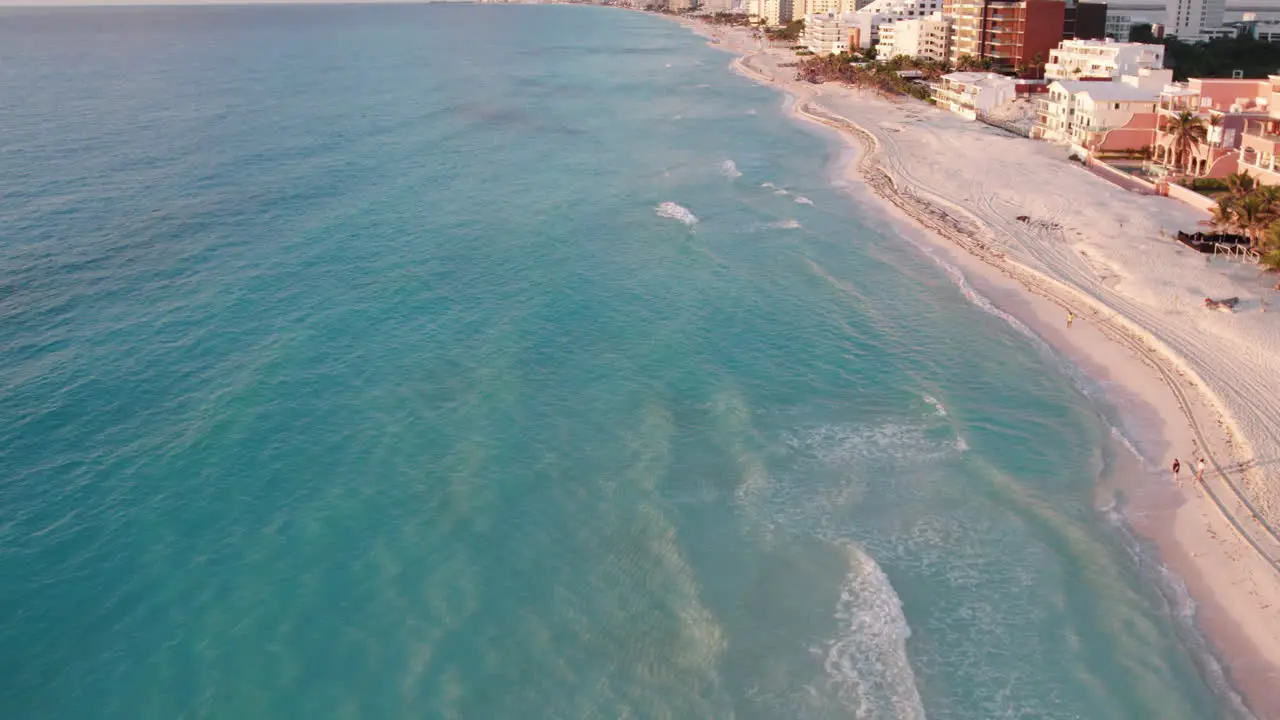 Aerial shot of rolling waves in a white sand beach surrounded by resorts and hotels in a crystal clear blue ocean in Cancun Mexico