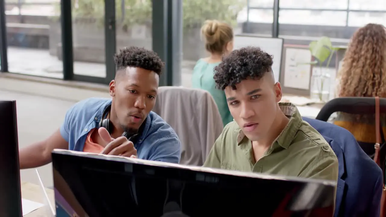 Two happy diverse male casual business colleagues in discussion at computer in office slow motion