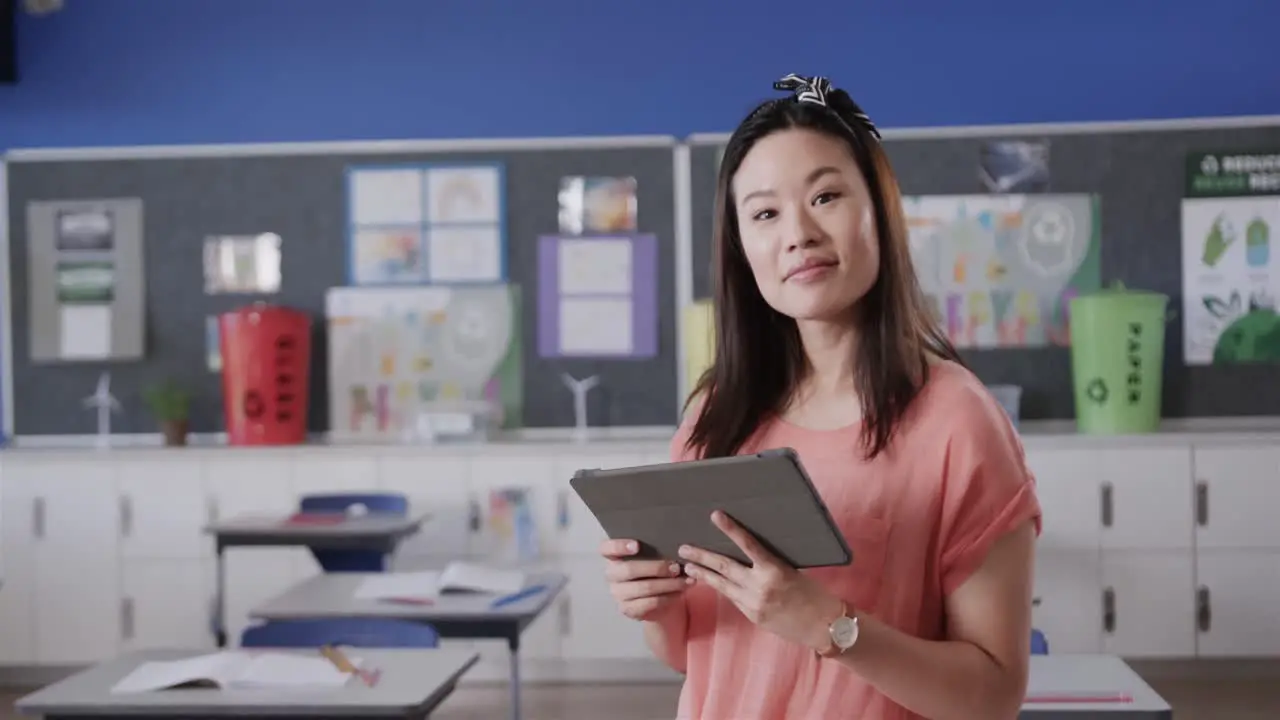 Portrait of happy asian female teacher with tablet in elementary school classroom slow motion