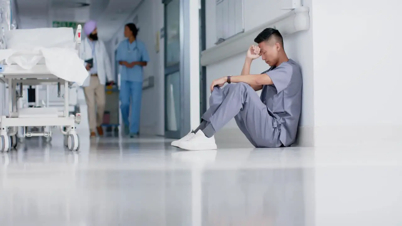 Stressed asian male surgeon sitting and leaning on wall in hospital in slow motion