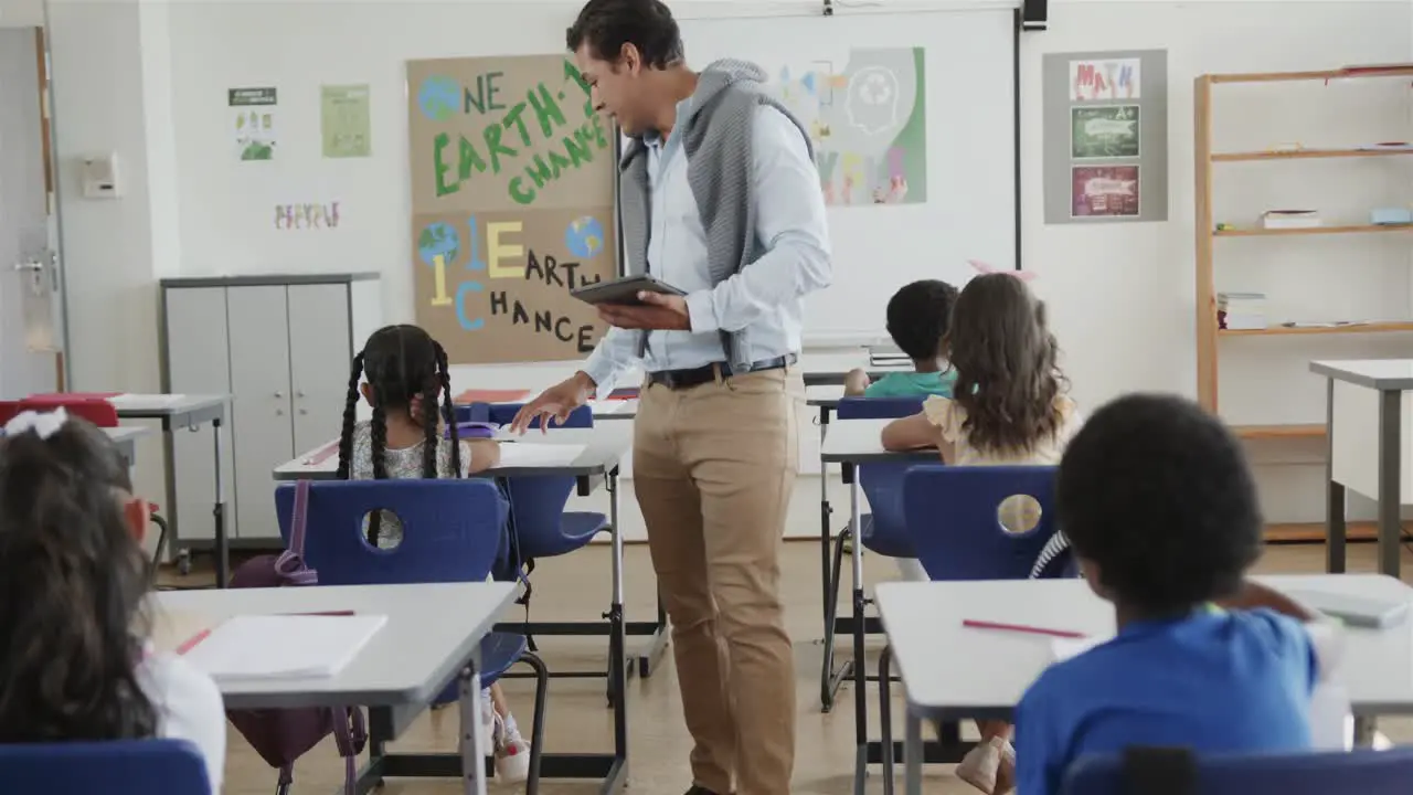 Diverse male teacher with tablet helping children at desks in elementary school class slow motion