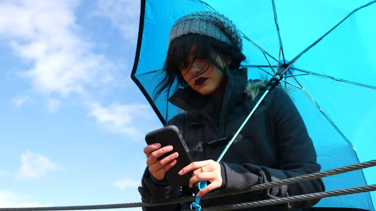 Attractive young woman texting on smartphone holding weather umbrella under blue skies as rain storm clouds pass