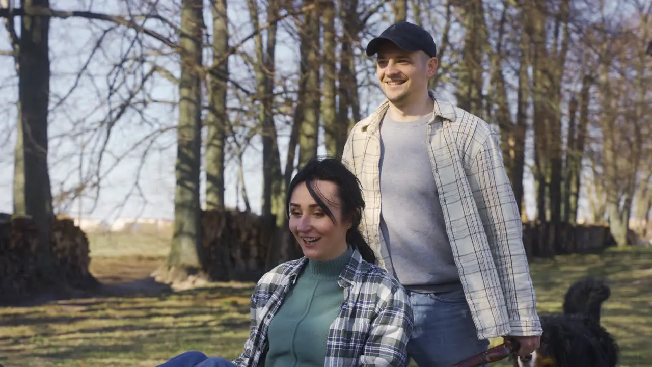 Caucasian man carrying his partner who has the arms opened on a wheelbarrow in the countryside Their dog walks around them