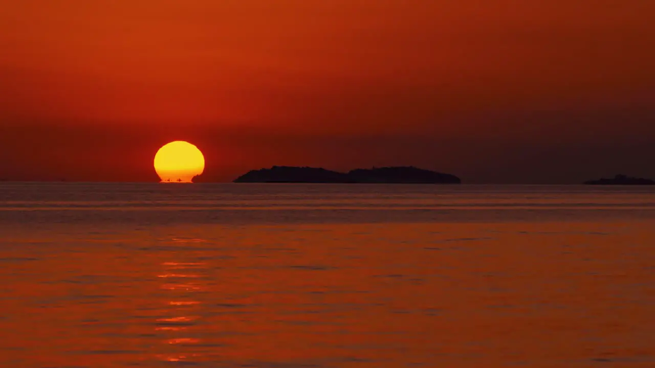 The sun is setting over the horizon at a romantic beach at the Croatian Mediterranean seaside as a big orange ball with sailing ships passing by