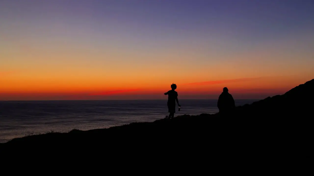 Silhouettes of adult walk along shoreline at sunset wide