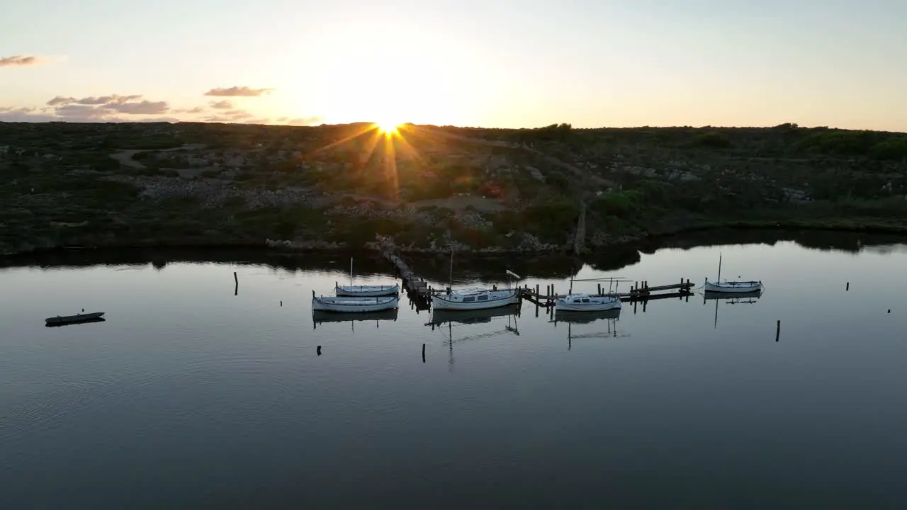 Aerial pullback above beautiful wooden harbor with sailboats docked in calm water as sun flare rises in sky