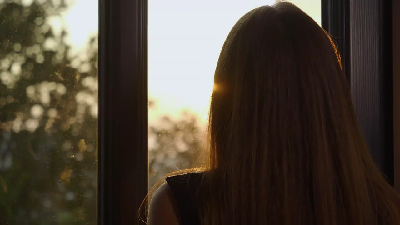 Medium handheld shot of a women looking out the window at the sunset and trees before turning to look into the camera while holding a glass of whiskey