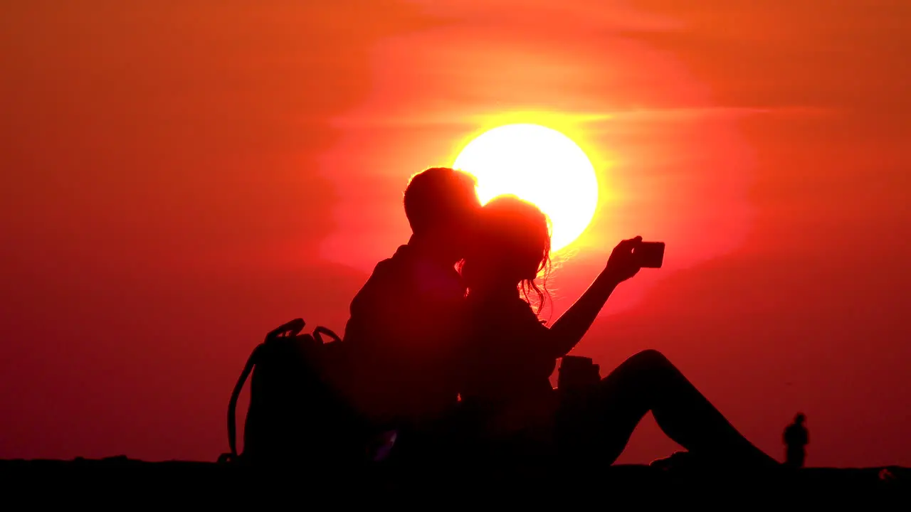 A couple sitting at North Avenue Beach in Chicago taking selfies on a cellphone in front of a bright yellow sun with orange sky