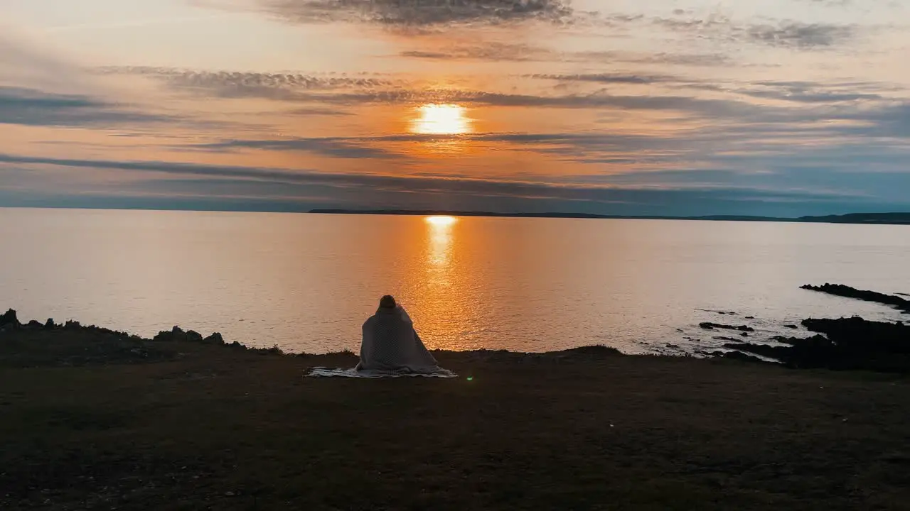 Beautiful young woman sitting on blanket on the top of mountain cliff in front of the Atlantic ocean at sunset