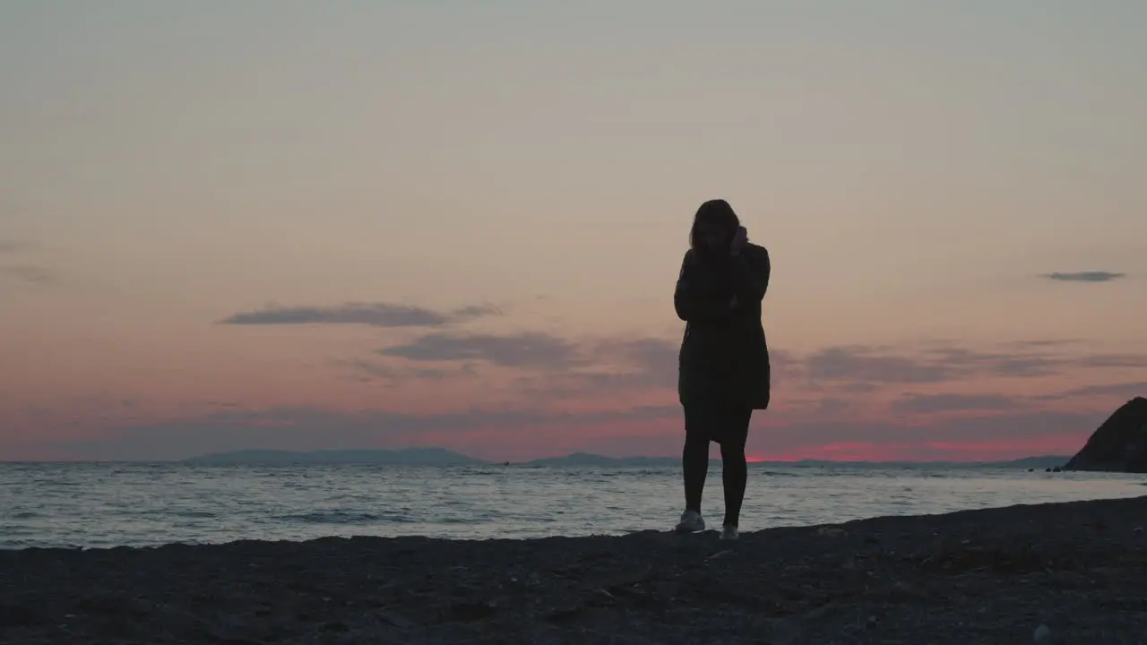 A girl wearing autumn jacket walking along the beach on the sunset with waves of the ocean washing ashore