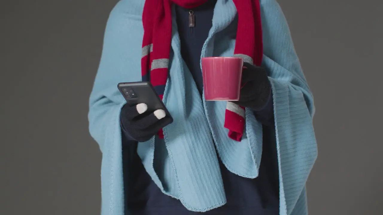 Close Up Of Person Wearing Blanket With Hot Drink Using Mobile Phone Trying To Keep Warm In Energy Crisis