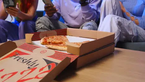 Close Up Of Gen Z Friends Sitting On Sofa Eating Home Delivery Takeaway Pizza And Using Mobile Phones