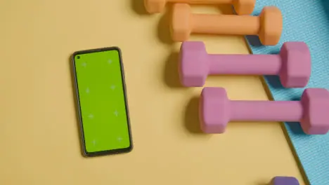 Overhead Fitness Studio Shot Of Hand Picking Up Coloured Exercise Dumbbell Weight Next To Green Screen Mobile Phone