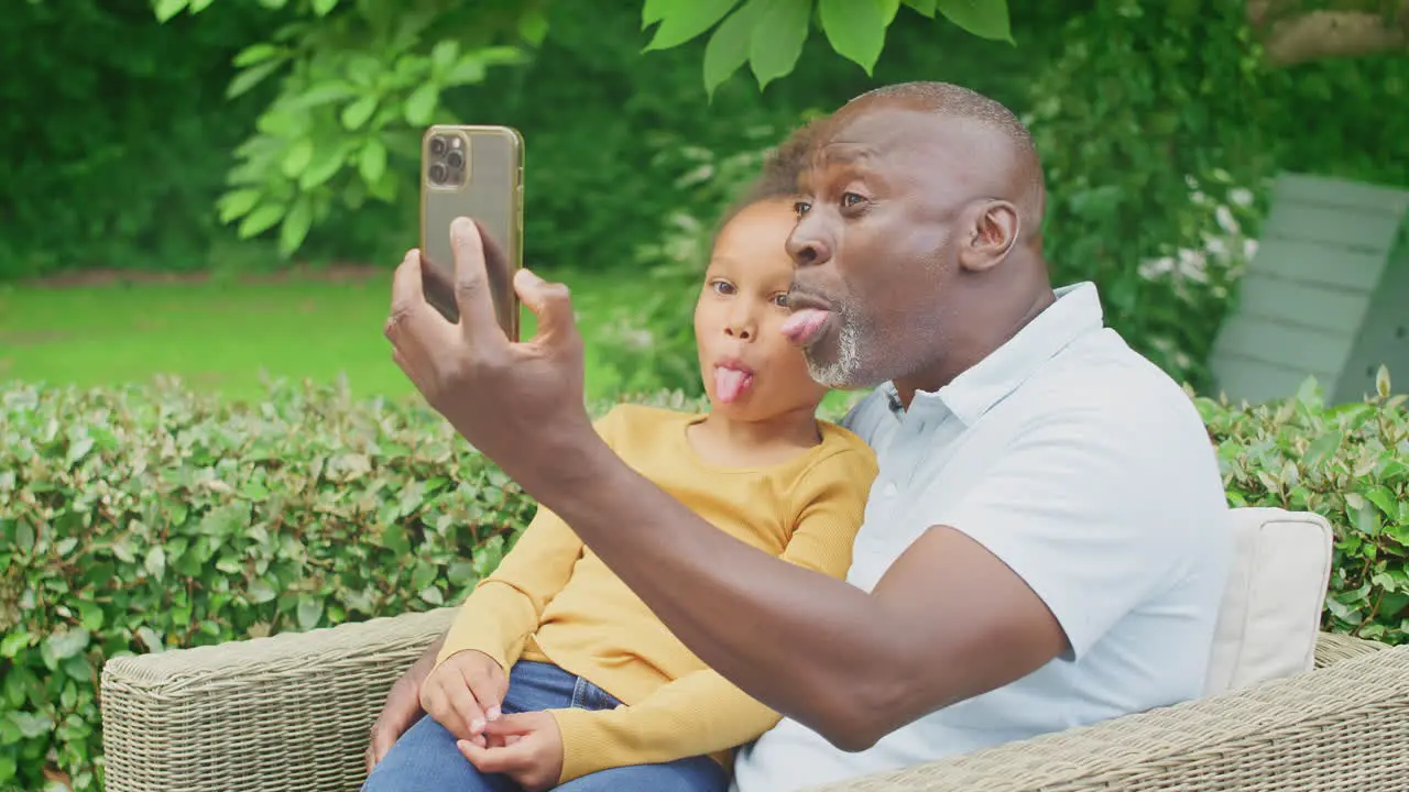 Grandfather Pulling Faces With Granddaughter Taking Selfie On Mobile Phone In Garden At Home