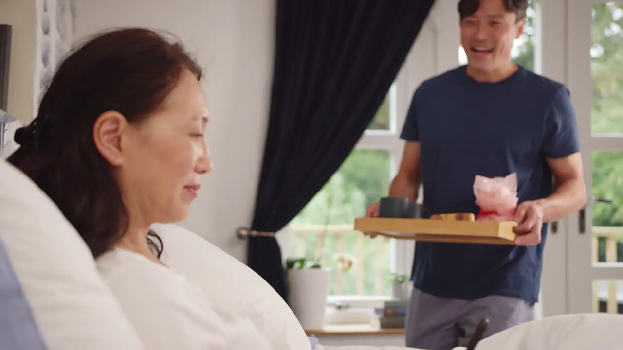 Mature Asian man bringing wife using mobile phone breakfast in bed to celebrate birthday or anniversary shot in slow motion