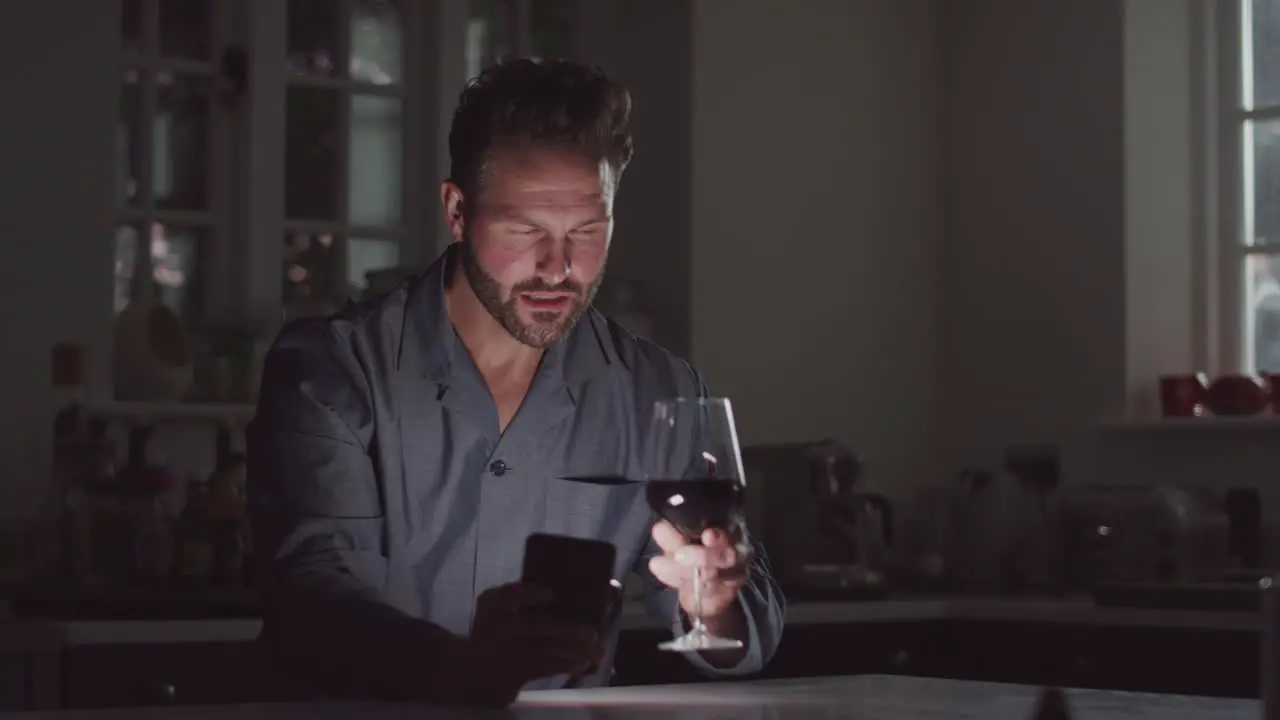 Mature Man Wearing Pyjamas Sitting In Kitchen With Glass Of Wine At Night Using Mobile Phone