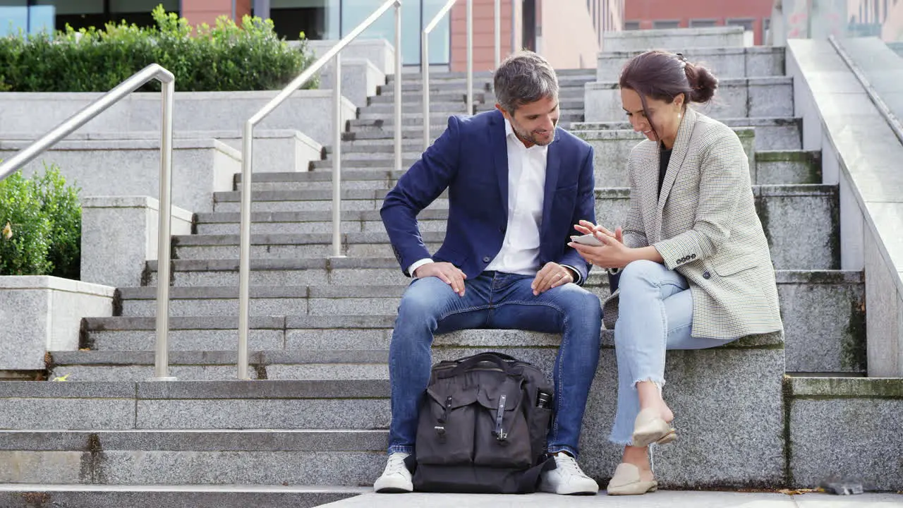 Businessman And Businesswoman Sitting By Steps Having Meeting Outdoors Looking At Phone