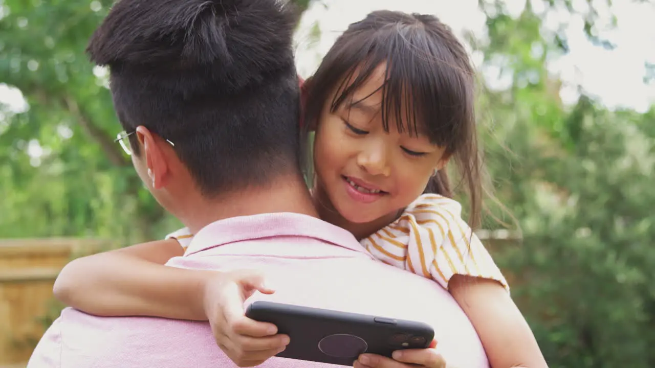 Asian Father Cuddling Girl In Garden As Girl Looks Over His Shoulder At Mobile Phone