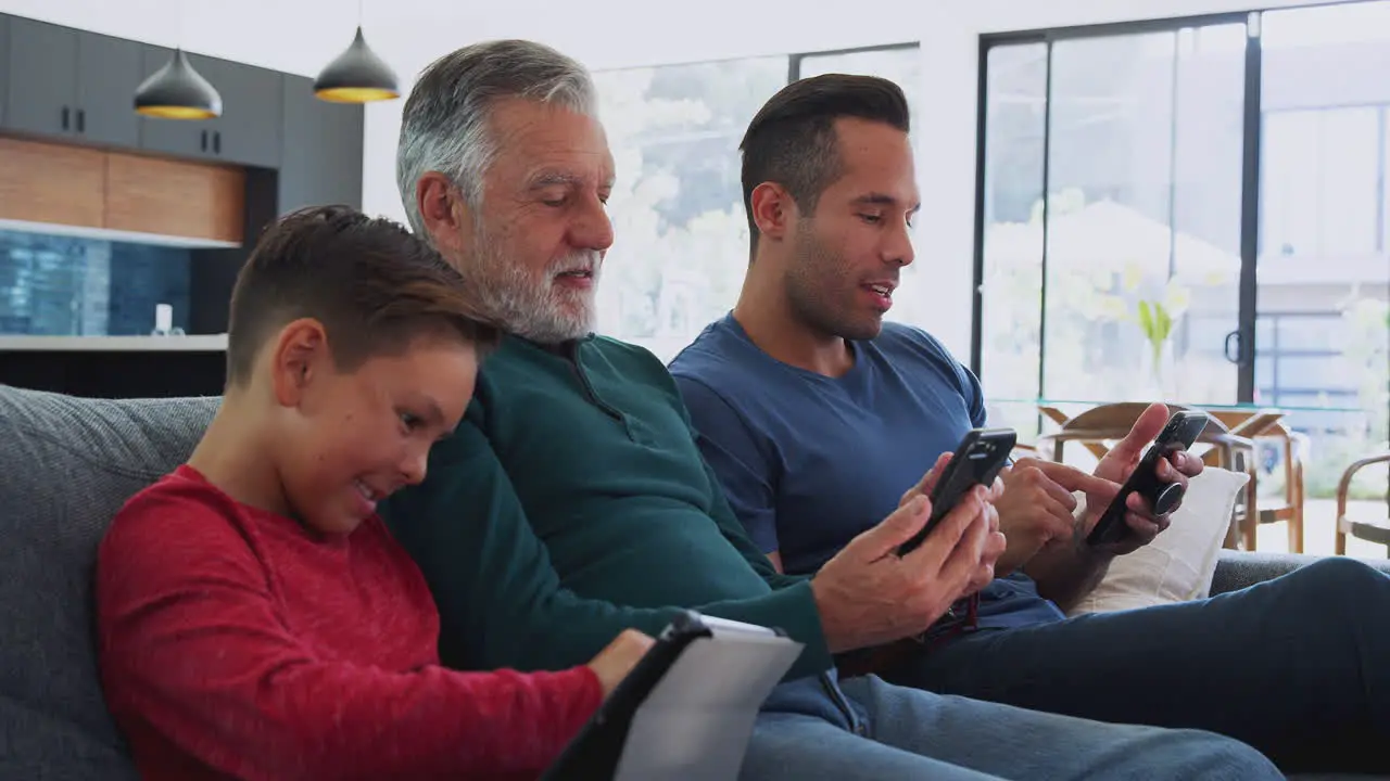 Multi-Generation Male Hispanic Family Sitting On Sofa At Home Using Mobile Phones And Digital Tablet