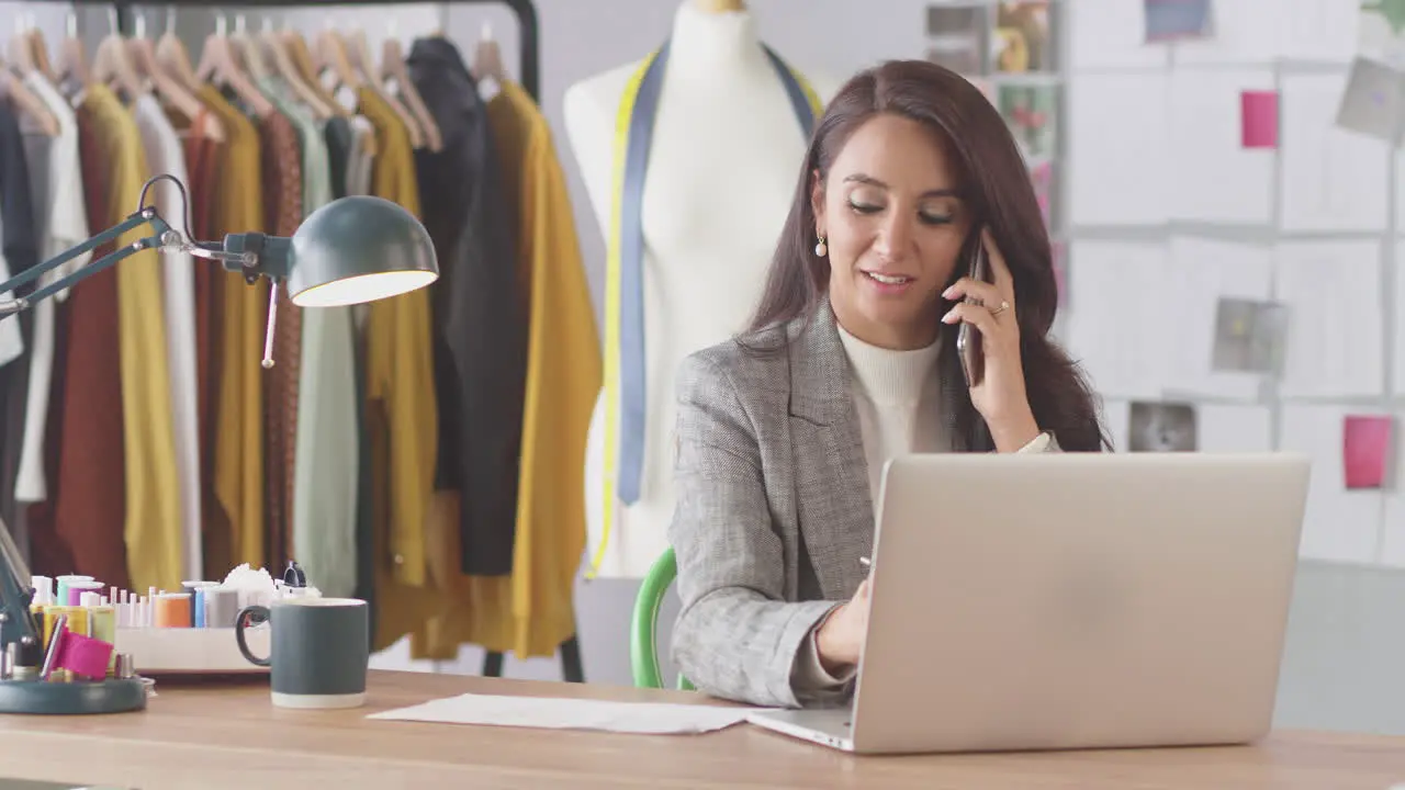 Female Fashion Designer In Studio Working At Desk On Laptop And Talking On Mobile Phone